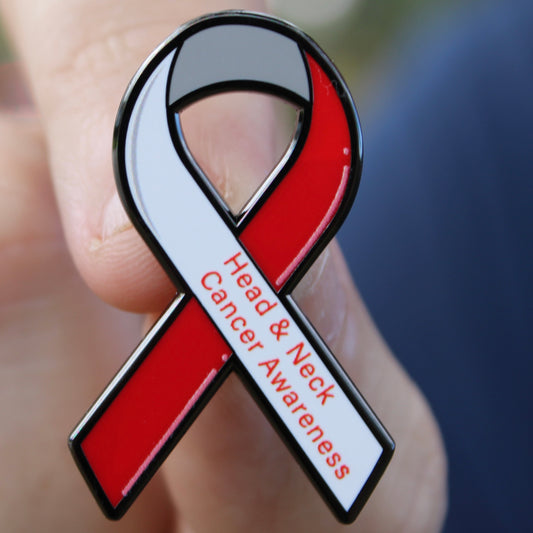 Red and white awareness ribbon pin for head and neck cancer held in a hand.