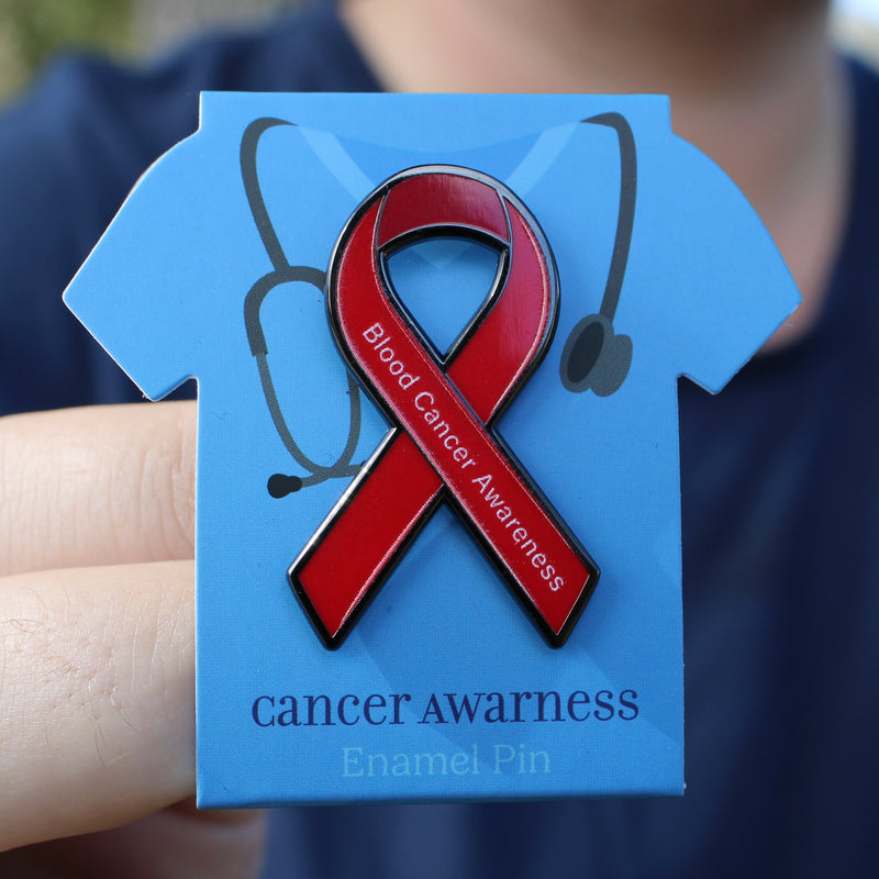 Red ribbon blood cancer awareness enamel pin on a blue card held by a hand.