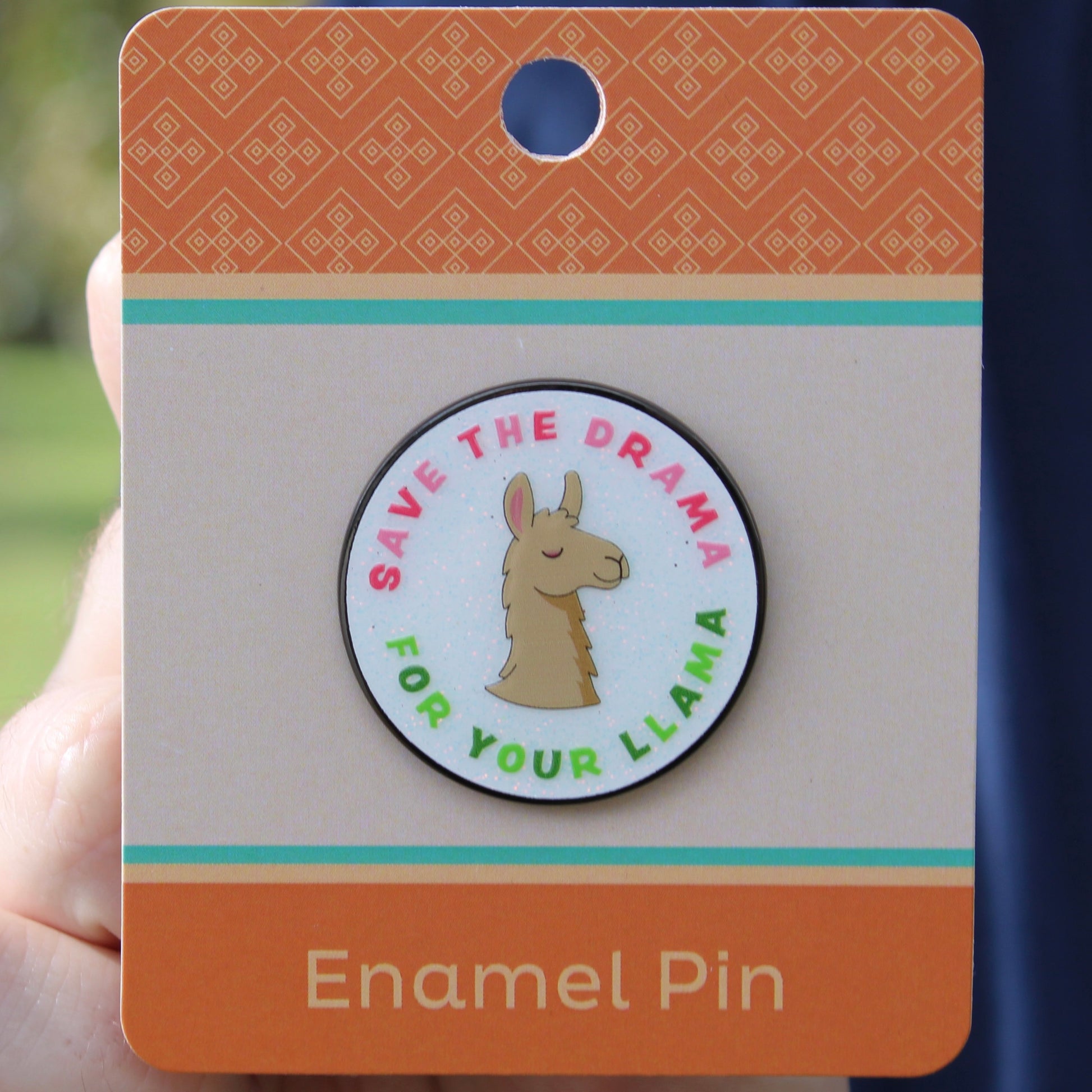 Enamel pin with font "save the drama for your llama" and a llama design on a card held by a hand