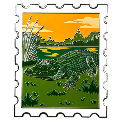 enamel pin shaped like a postage stamp featuring an american alligator at sunset in the everglades